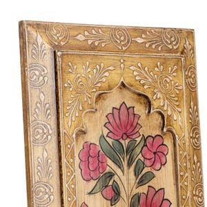 May include: Close-up of a decorative wooden panel with a gold-coloured frame and floral design. The panel features a central floral motif with pink flowers and green leaves, set against a light-coloured background. Intricate white carvings adorn the frame.