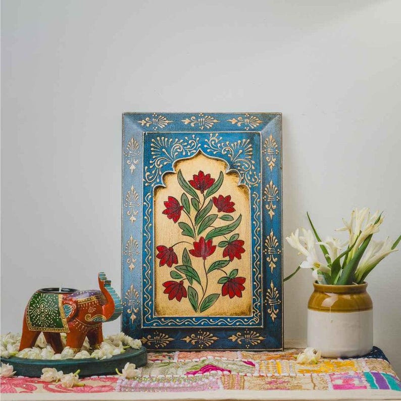 May include: A blue wooden framed artwork with a gold floral design. The artwork features a central floral motif with red flowers and green leaves. An elephant candle holder and a vase with white flowers are also present.