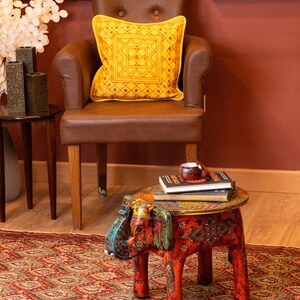May include: A yellow decorative pillow with a geometric design sits on a brown leather armchair. A small, red, elephant-shaped table with colorful patterns holds books and a teacup. The room has a red rug and walls.