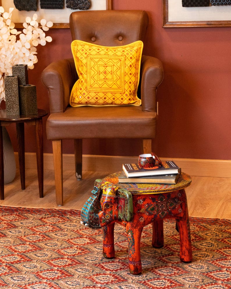May include: A yellow decorative pillow with a geometric design sits on a brown leather armchair. A small, red, elephant-shaped table with colorful patterns holds books and a teacup. The room has a red rug and walls.