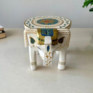 May include: A white, hand-painted wooden elephant stool with intricate gold, blue, and teal details. The round top features a floral design, and the elephant's head has a gold and blue decorative piece. The legs are painted with gold accents.