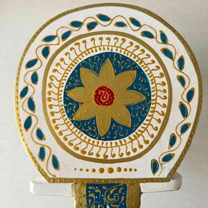 May include: A decorative, hand-painted wooden piece featuring a circular design. The central element is a gold flower with a red center, set against a blue background. The design is framed by gold and blue accents on a white base.