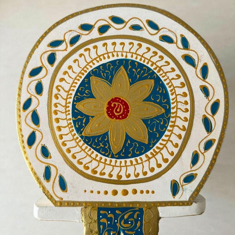 May include: A decorative, hand-painted wooden piece featuring a circular design. The central element is a gold flower with a red center, set against a blue background. The design is framed by gold and blue accents on a white base.