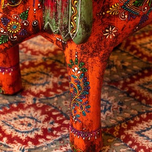 May include: Close-up of a handcrafted, orange wooden elephant figurine with intricate floral and paisley designs in various colors. The elephant's legs and body are adorned with detailed patterns, and the saddle is green.