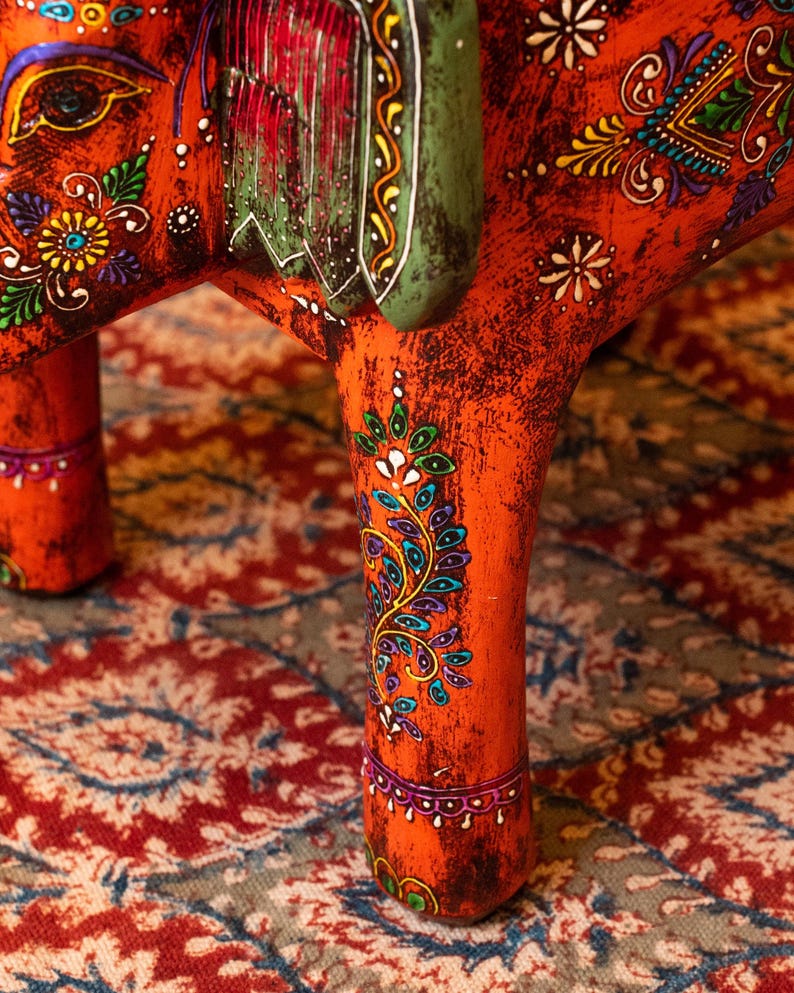 May include: Close-up of a handcrafted, orange wooden elephant figurine with intricate floral and paisley designs in various colors. The elephant's legs and body are adorned with detailed patterns, and the saddle is green.