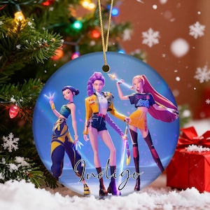 May include: A round Christmas ornament featuring three stylized female figures in fashionable outfits. The ornament has a blue background with the word "Indigo" in a script font. The ornament is hanging from a gold string.