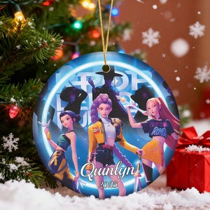May include: A round Christmas ornament with a gold string. The ornament features a group of stylized figures in a blue and white design. The name "Quinlyn" and the year "2025" are printed on the ornament.