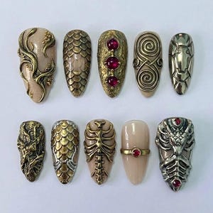 May include: A collection of ten ornate, sculpted nail designs. The nails feature a variety of textures and embellishments, including metallic accents, scale patterns, and embedded red gemstones. The designs showcase a blend of artistic and gothic elements.
