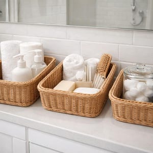 May include: Three woven wicker baskets on a white countertop, each filled with bathroom essentials. The baskets contain white towels, soap, cotton balls, and toiletries. A small potted plant is visible in the background.