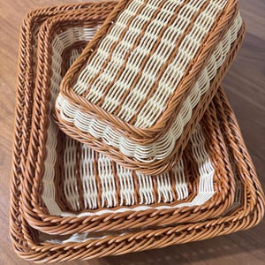 May include: A set of three rectangular woven baskets featuring a two-tone design with light cream and medium brown strands. The baskets are nested together, each displaying a braided rim and a vertical weave pattern on the sides and base.