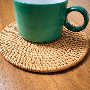 May include: A green ceramic mug with a large handle sits on a round, woven rattan coaster. The mug has a white interior and a glossy exterior. The coaster is made of natural-colored woven material. The mug and coaster are on a wooden surface.