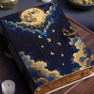May include: A decorative journal with a celestial design featuring a full moon, clouds, and crescent moons. The cover is dark blue with gold accents and a metal clasp. The journal is on a wooden tray with candles and small jars.