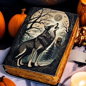 May include: A decorative journal with a wolf howling at the moon design. The cover is dark with a detailed illustration of a wolf, trees, and a full moon. The journal has a clasp closure and aged-looking pages. Pumpkins and candles surround the journal.