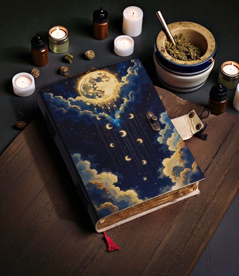 May include: A decorative journal with a celestial design featuring a full moon, crescent moons, and clouds. The cover is dark blue with gold accents. A red tassel marks the pages. Candles and small bottles surround the journal.