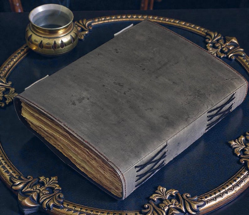 Puede incluir: Un libro gris encuadernado en cuero con costuras visibles y p&aacute;ginas envejecidas. El libro est&aacute; sobre una superficie azul oscuro, junto a un marco decorativo dorado y un peque&ntilde;o recipiente de metal adornado.