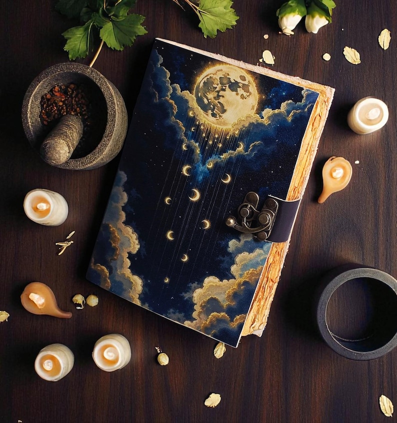 May include: A journal with a celestial design featuring a full moon, crescent moons, and clouds. The cover is dark blue with gold and white accents. The journal is open, revealing aged paper. Small candles and a mortar and pestle surround the journal.