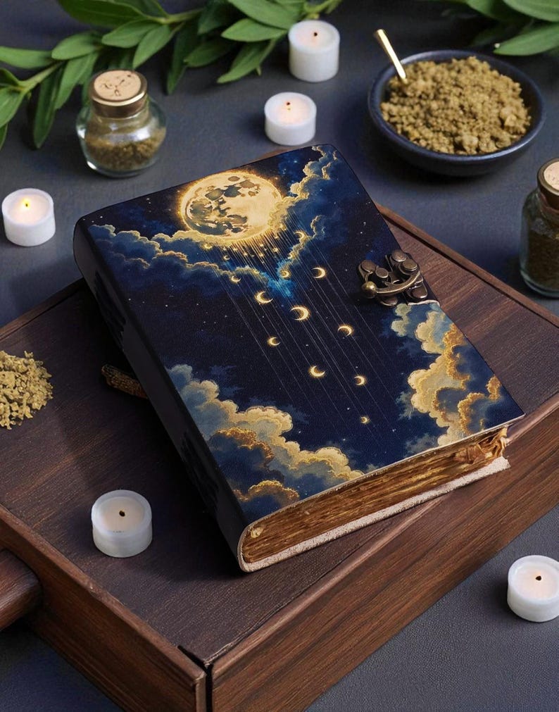 May include: A decorative journal with a celestial design featuring a full moon, clouds, and crescent moons. The cover is dark blue with gold accents and a metal clasp. The journal is on a wooden tray with candles and small jars.