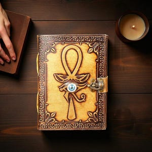 May include: A leather journal with an embossed Ankh symbol and Eye of Horus design. The journal has a brass clasp and a blue eye detail. The cover is tan with brown decorative borders. A lit candle sits nearby.