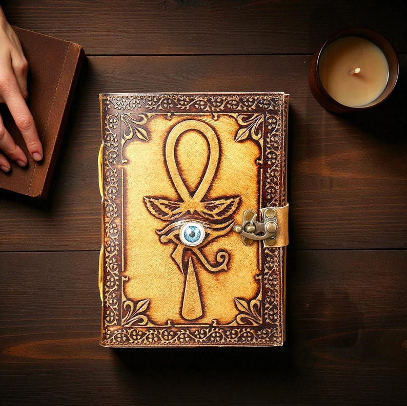 May include: A leather journal with an embossed Ankh symbol and Eye of Horus design. The journal has a brass clasp and a blue eye detail. The cover is tan with brown decorative borders. A lit candle sits nearby.