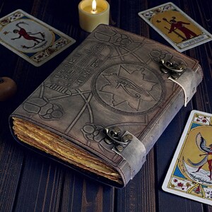 Puede incluir: Un diario de cuero marr&oacute;n desgastado con dise&ntilde;os egipcios en relieve, incluyendo escarabajos y jerogl&iacute;ficos. El diario tiene un cierre de metal y p&aacute;ginas envejecidas y amarillentas. Cartas del tarot y una vela encendida est&aacute;n dispuestas alrededor del libro.