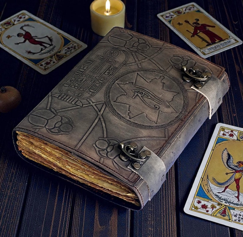 Puede incluir: Un diario de cuero marr&oacute;n desgastado con dise&ntilde;os egipcios en relieve, incluyendo escarabajos y jerogl&iacute;ficos. El diario tiene un cierre de metal y p&aacute;ginas envejecidas y amarillentas. Cartas del tarot y una vela encendida est&aacute;n dispuestas alrededor del libro.