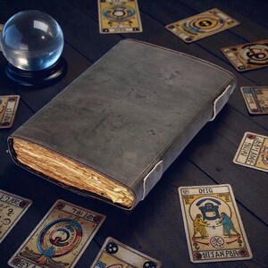 Puede incluir: Un libro grande, gris y desgastado con p&aacute;ginas amarillentas, acompa&ntilde;ado de cartas del tarot y una bola de cristal. El libro tiene una cubierta de cuero y cierres blancos. Las cartas del tarot presentan ilustraciones y texto.