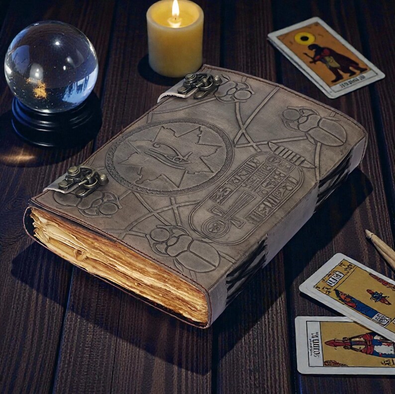 Puede incluir: Un diario de cuero gris desgastado con jerogl&iacute;ficos y s&iacute;mbolos egipcios en relieve. El diario est&aacute; asegurado con un cierre de metal y se encuentra sobre una superficie de madera oscura, acompa&ntilde;ado de cartas del tarot, una bola de cristal y una vela encendida.