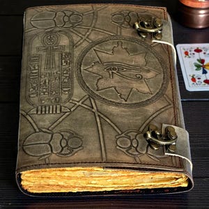 Puede incluir: Un diario de cuero marr&oacute;n con s&iacute;mbolos egipcios en relieve, incluyendo un ankh, el ojo de Horus y escarabajos. El diario tiene un cierre de hebilla y p&aacute;ginas amarillentas. La cubierta est&aacute; detallada con patrones geom&eacute;tricos.