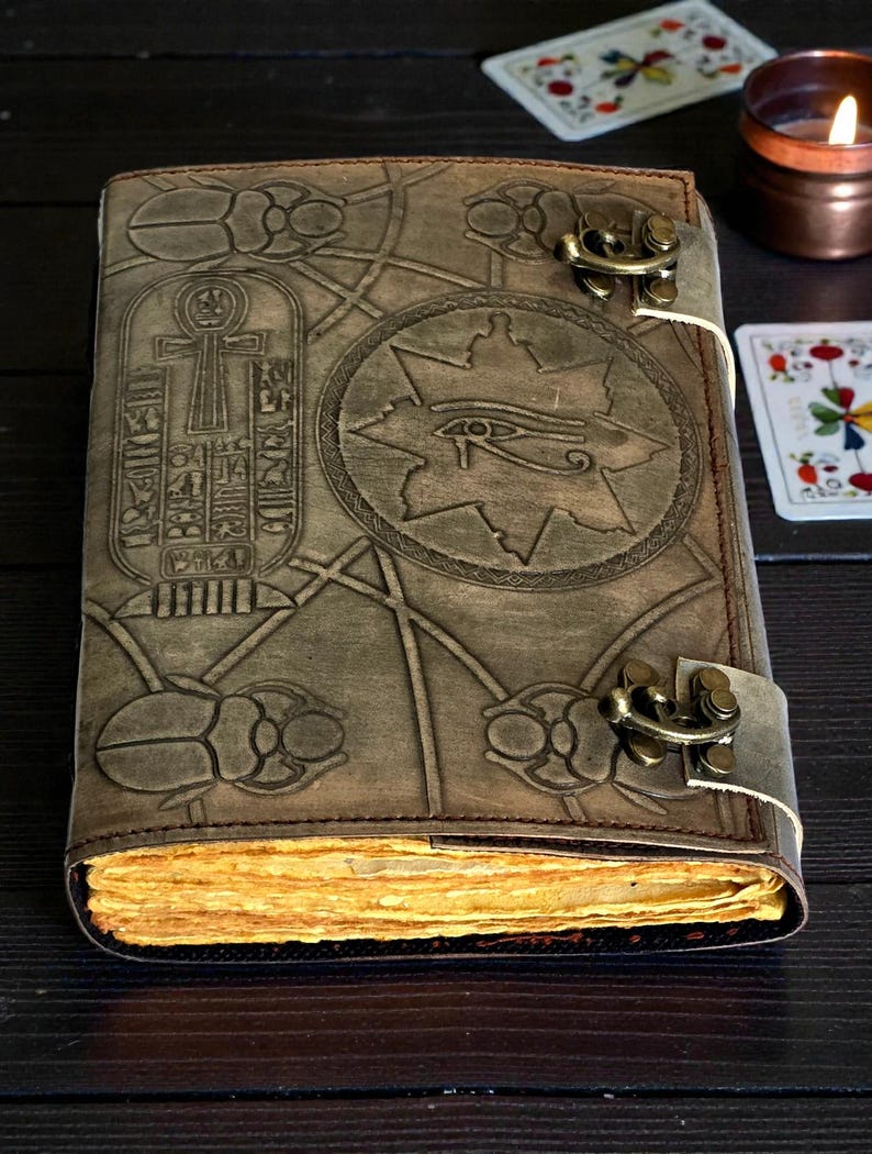 Puede incluir: Un diario de cuero marr&oacute;n con s&iacute;mbolos egipcios en relieve, incluyendo un ankh, el ojo de Horus y escarabajos. El diario tiene un cierre de hebilla y p&aacute;ginas amarillentas. La cubierta est&aacute; detallada con patrones geom&eacute;tricos.