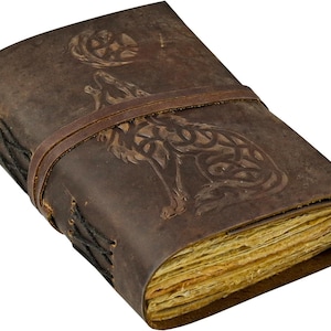 May include: A brown leather journal with a wolf howling at the moon design embossed on the cover. The journal is secured with a leather strap and features aged, yellowed pages. The edges are stitched with black thread.