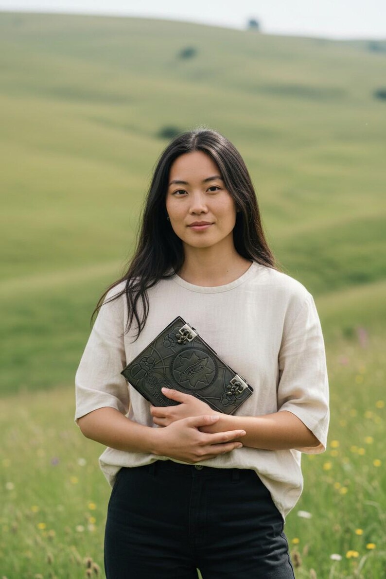 Puede incluir: Un diario de cuero verde oscuro con intrincados dise&ntilde;os en relieve y cierres met&aacute;licos. El diario es sostenido por una persona que lleva una camisa color crema y pantalones negros, de pie en un campo verde.