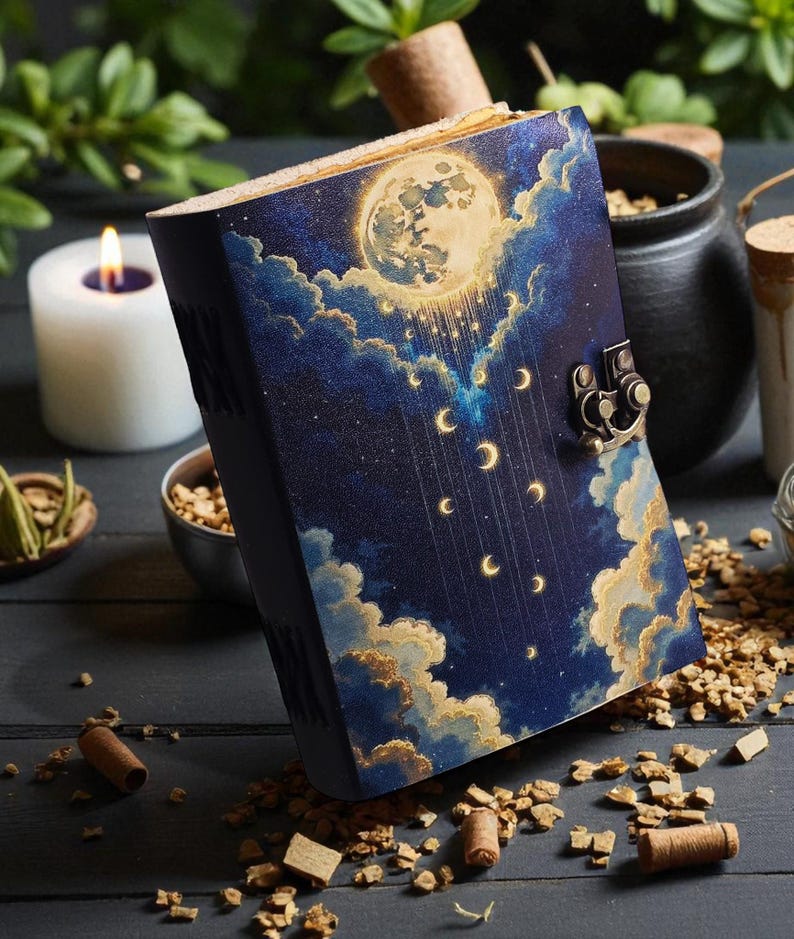 May include: A hardcover journal with a celestial design featuring a full moon, clouds, and crescent moons against a dark blue background. The journal has a metal clasp and is surrounded by candles and herbs.