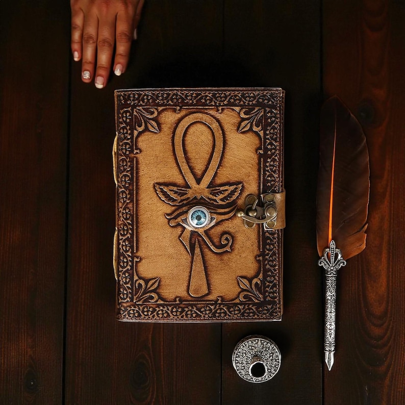 May include: A brown leather journal featuring an embossed ankh symbol and a blue eye detail. A metal clasp secures the journal, which is displayed with a feather pen and a small, round inkwell. The items are on a dark wooden surface.