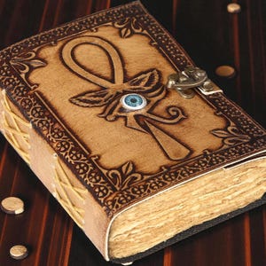 May include: A tan leather journal with an embossed ankh symbol and a blue eye detail. The cover features decorative borders and a metal clasp. The pages are aged, and the spine is stitched with tan thread.