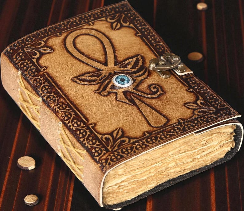 May include: A tan leather journal with an embossed ankh symbol and a blue eye detail. The cover features decorative borders and a metal clasp. The pages are aged, and the spine is stitched with tan thread.