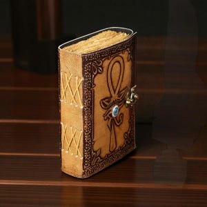 May include: A leather journal with an ankh symbol and clasp. The tan cover has brown embossed details and stitched edges. The journal has aged, yellowed pages and a blue gem. The journal is likely made of genuine leather.