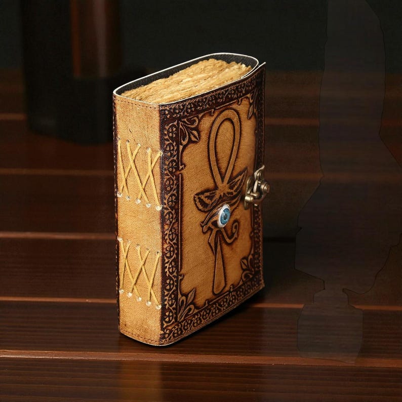 May include: A leather journal with an ankh symbol and clasp. The tan cover has brown embossed details and stitched edges. The journal has aged, yellowed pages and a blue gem. The journal is likely made of genuine leather.