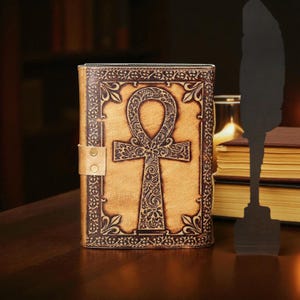 May include: A tan leather journal featuring a dark brown embossed ankh design. The journal has a decorative border and a snap closure. The background includes a stack of books and a quill silhouette.