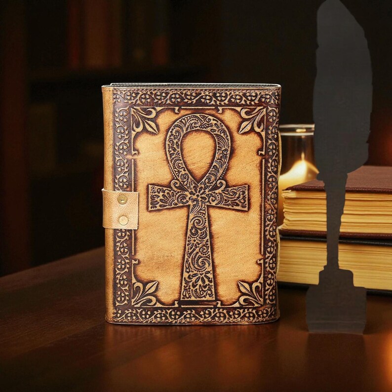 May include: A tan leather journal featuring a dark brown embossed ankh design. The journal has a decorative border and a snap closure. The background includes a stack of books and a quill silhouette.