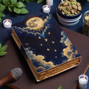 May include: A hardcover book with a celestial design featuring a full moon, crescent moons, and clouds. The cover is dark blue with gold and white accents. The aged pages are visible. The book is on a brown surface with candles and other items.