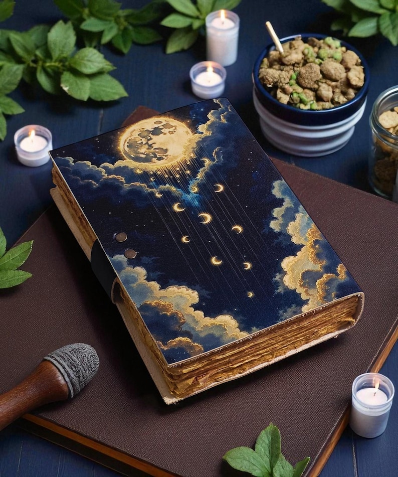 May include: A hardcover book with a celestial design featuring a full moon, crescent moons, and clouds. The cover is dark blue with gold and white accents. The aged pages are visible. The book is on a brown surface with candles and other items.