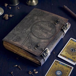 Puede incluir: Un diario de cuero gris con intrincados dise&ntilde;os en relieve, incluyendo jerogl&iacute;ficos y un s&iacute;mbolo de ojo. El diario est&aacute; asegurado con cierres met&aacute;licos y est&aacute; abierto, revelando p&aacute;ginas envejecidas y amarillentas. Se ven dos cartas de tarot.