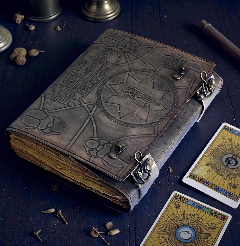 Puede incluir: Un diario de cuero gris con intrincados dise&ntilde;os en relieve, incluyendo jerogl&iacute;ficos y un s&iacute;mbolo de ojo. El diario est&aacute; asegurado con cierres met&aacute;licos y est&aacute; abierto, revelando p&aacute;ginas envejecidas y amarillentas. Se ven dos cartas de tarot.