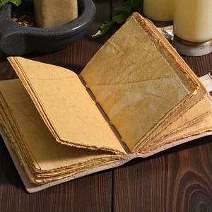 May include: An open, antique-style journal with aged, yellowed pages. The book is bound with a light-colored cover and held open with a metal clip. The pages have a textured, handmade appearance, suggesting a vintage or handcrafted item.