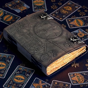 Puede incluir: Un diario de cuero gris oscuro con intrincados dise&ntilde;os en relieve, incluyendo un s&iacute;mbolo de ojo y jerogl&iacute;ficos. El diario est&aacute; asegurado con cierres met&aacute;licos y rodeado de cartas de tarot sobre una superficie azul oscuro. Las p&aacute;ginas tienen una apariencia c&aacute;lida y envejecida.