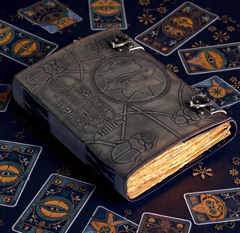 Puede incluir: Un diario de cuero gris oscuro con intrincados dise&ntilde;os en relieve, incluyendo un s&iacute;mbolo de ojo y jerogl&iacute;ficos. El diario est&aacute; asegurado con cierres met&aacute;licos y rodeado de cartas de tarot sobre una superficie azul oscuro. Las p&aacute;ginas tienen una apariencia c&aacute;lida y envejecida.