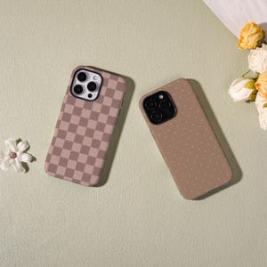 May include: Two phone cases with different designs. One case has a brown and tan checkerboard pattern. The other case is a solid brown color with small white polka dots. Both cases have a black camera lens cutout.