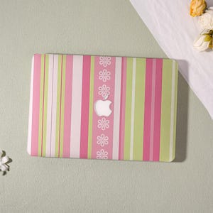 May include: A laptop skin featuring a vertical striped pattern in shades of pink, light green, and white. A central vertical band displays a repeating white flower motif, with a cutout in the middle revealing the silver Apple logo.