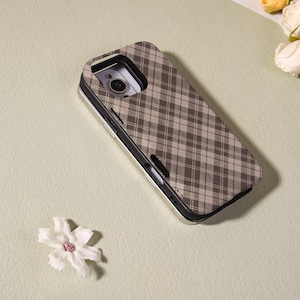 May include: A phone case with a brown and beige plaid pattern. The case has a metallic gold-colored frame and a camera cutout. A white flower with a pink center is in the foreground.