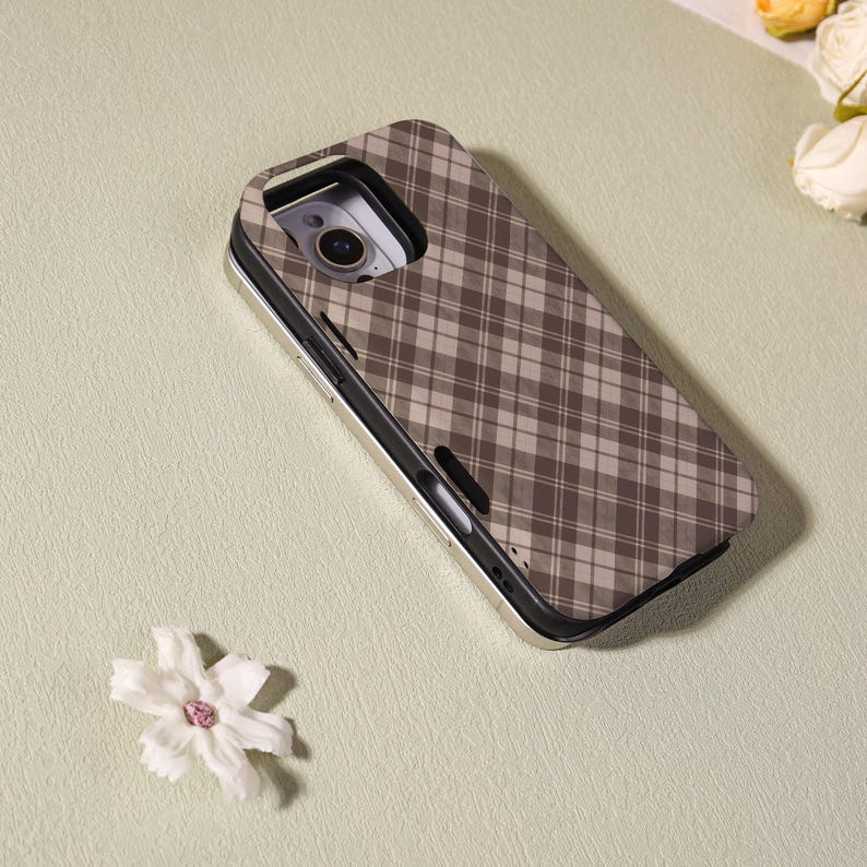 May include: A phone case with a brown and beige plaid pattern. The case has a metallic gold-colored frame and a camera cutout. A white flower with a pink center is in the foreground.